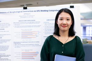 Winner of the popular vote category Jiefang Li smiles in front of her academic poster at the inaugural Thomas M. Carsey Graduate Student Symposium. The poster includes blue text on white background with two horizontal bar charts and a cut off title, "...iveness of Morphological Instruction on EFL Reading Comprehension." Jiefang Li wears a dark green blouse and holds a blue folder.