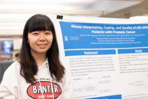 Student smiles in front of a research poster. A partially visible title reads "Illness uncertainty, coping, and quality of life among... patients with prostate cancer."