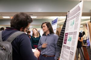 A student speaks about their research to attendees in front of a research poster, the text of which is not readable.