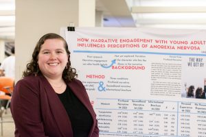 Student smiles in front of a research poster. A partial title is visible: "How narrative engagement with young adult... influences perceptions of anorexia nervosa."