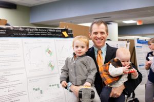 Student smiles in front of a research poster with two children in their hands. The poster contains several graphs and charts, and is titled "Lexical entropy quantifies discourse production severity."