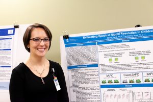 Student in front of a research poster, featuring visible abstract, background, methods, and results sections, entitled "Estimating Spectral Ripple Resolution in Children..." The full title is cut off.
