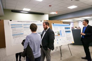 Student presents their research to another individual in front of a research poster.