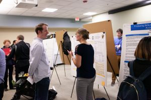 Student presents their research to another individual in front of a research poster.